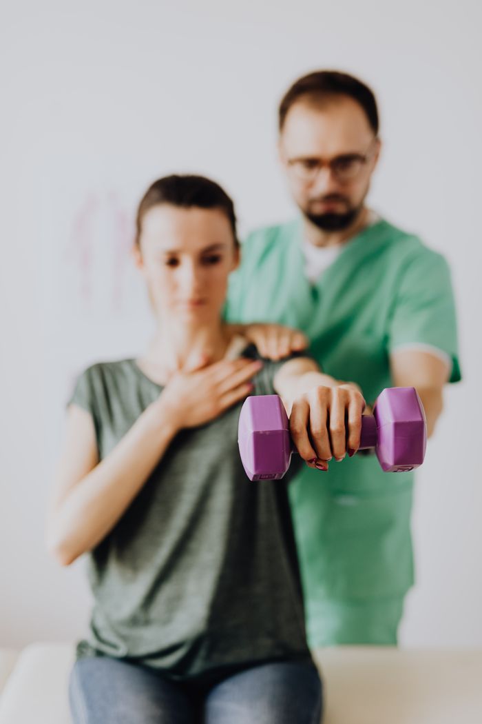 Bearded orthopedist in uniform and eyeglasses helping slim woman in casual clothes reaching arm with dumbbell while sitting on examination couch in clinic